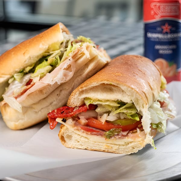 Two Italian deli sandwiches with various fillings, lettuce, and tomatoes, next to a San Pellegrino can.